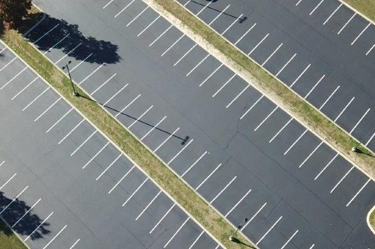 Aerial view of a mostly empty asphalt parking lot with white parking space lines. Green medians separate rows.