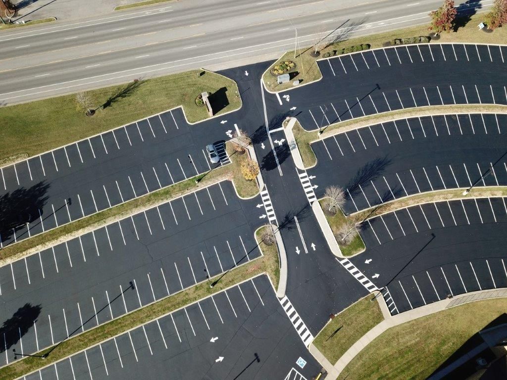 Aerial view of a white, L-shaped building with surrounding black asphalt parking and road, nestled in green trees and grass.