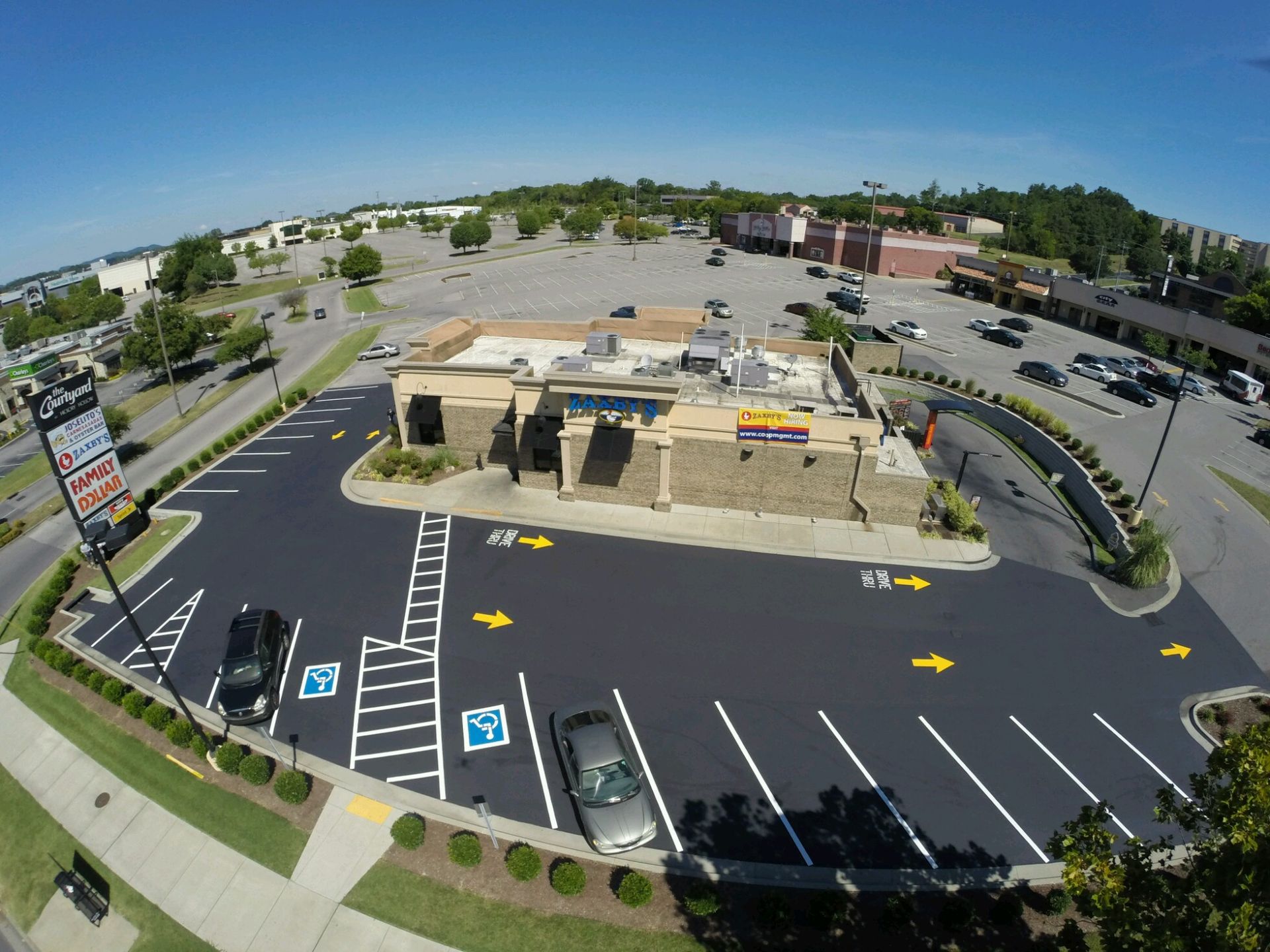 Aerial view of a white, L-shaped building with surrounding black asphalt parking and road, nestled in green trees and grass.