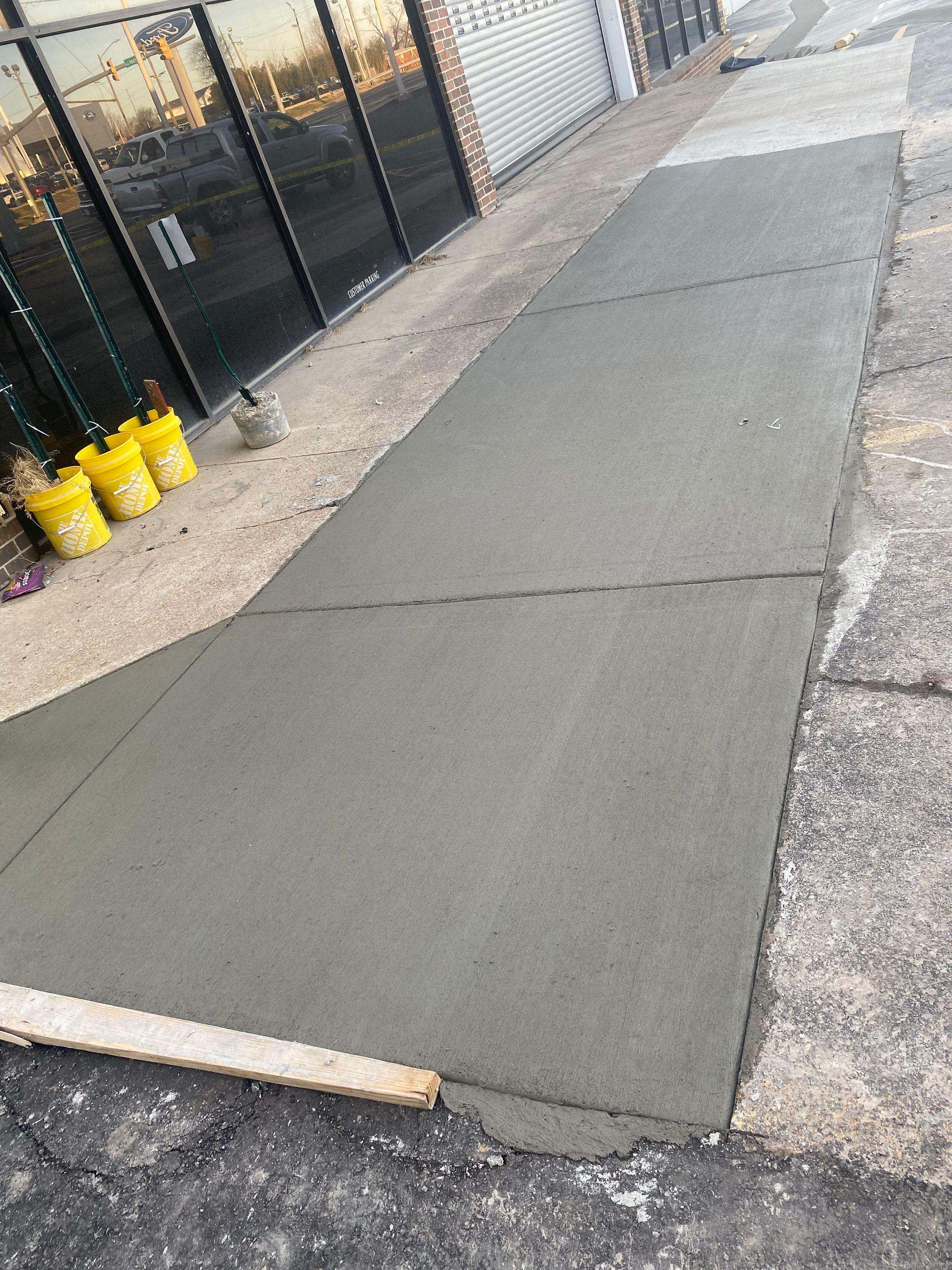 Freshly poured concrete sidewalk next to a building. Wooden straightedge lies at the bottom.