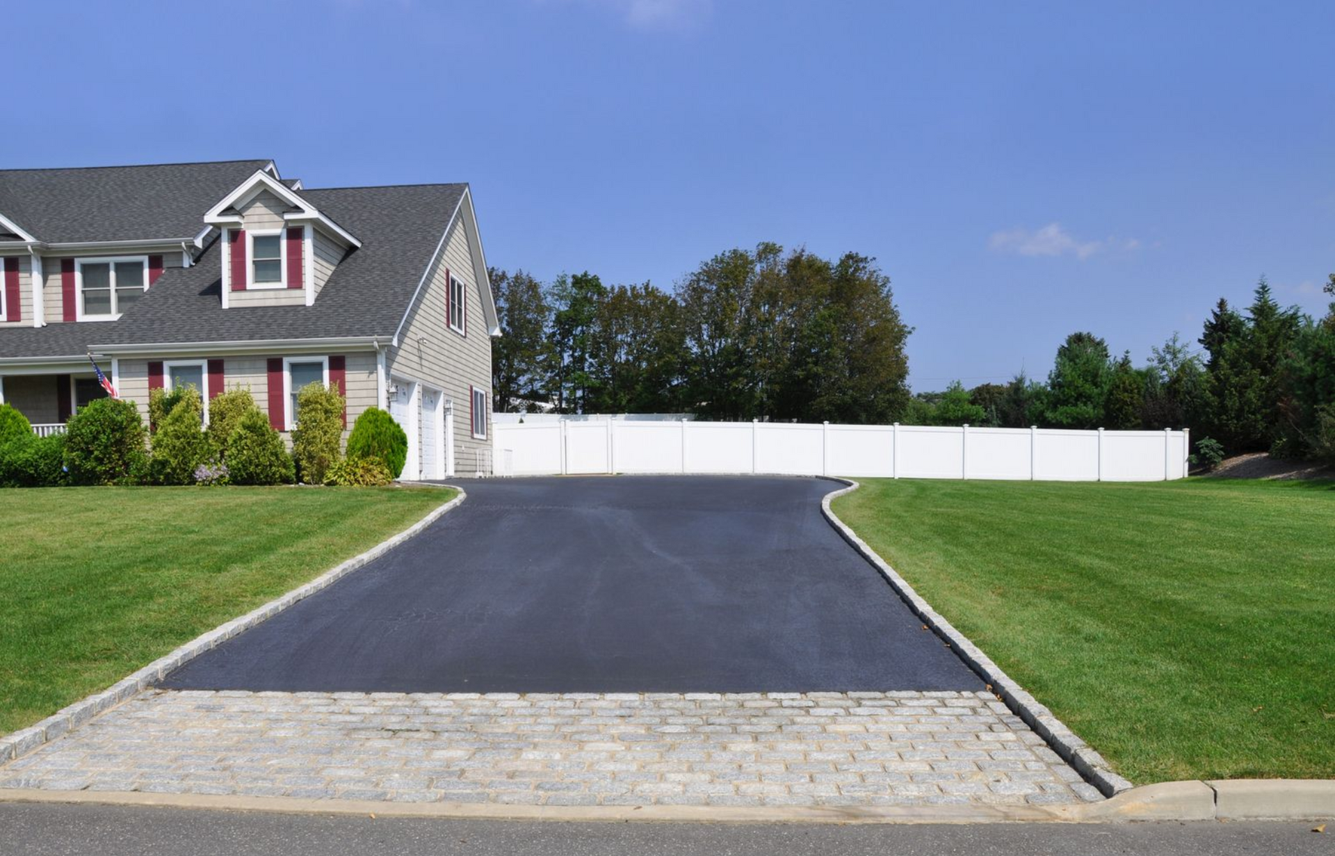A house with a black asphalt driveway, green lawn, and white fence under a blue sky.