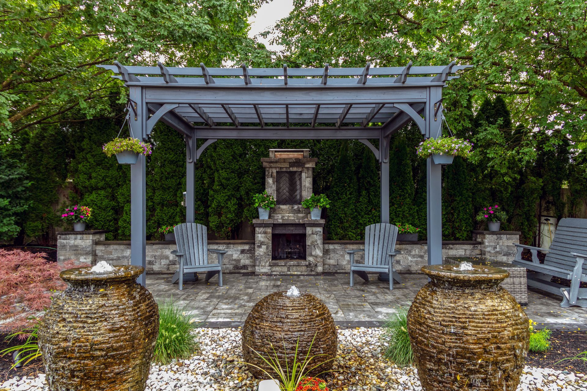 Gray pergola with fireplace, seating, and water fountains in a stone patio garden.