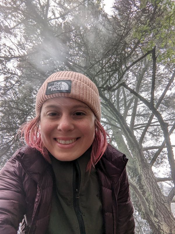 A woman wearing a beanie and a jacket is smiling in front of a tree.