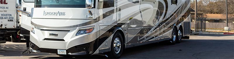 Large silver and black RV parked on pavement next to a white truck. Day, exterior.