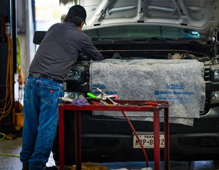 Mechanic working on a car engine in a garage; tools on a red cart. Texas license plate visible.
