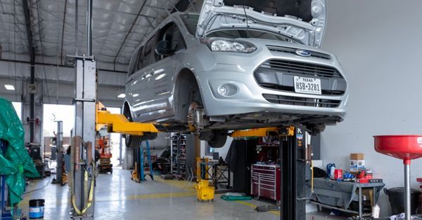 Silver Ford van on a lift in a repair shop, hood open, wheels removed.