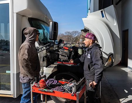 Two people working on a truck engine in an outdoor setting. The hood is open.