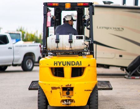Yellow Hyundai forklift, propane tank, driver, RV, outside.