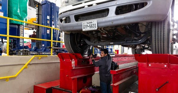 Mechanic working under a raised truck in a repair shop; red lift, gray truck, Texas license plate.