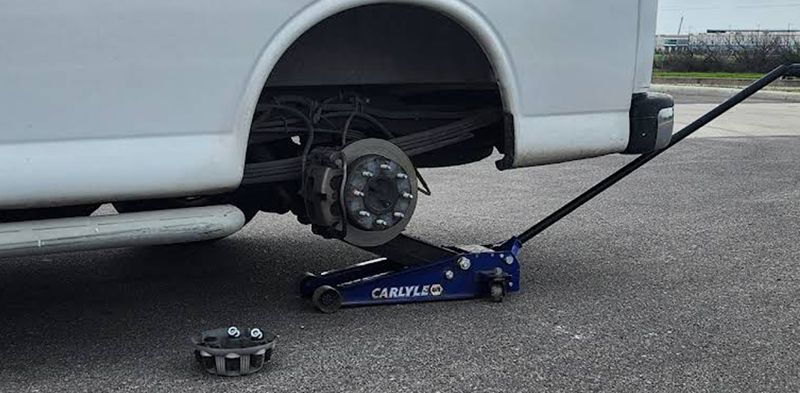 A white van with a flat tire being lifted by a blue jack. A wheel sits on the ground.
