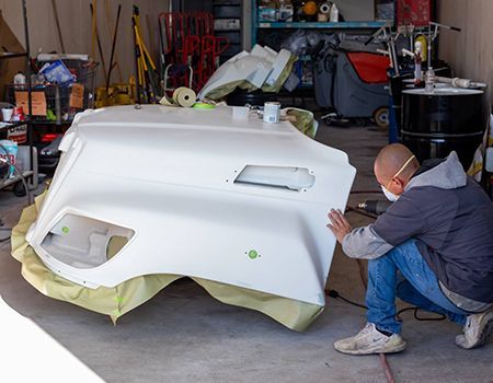 Person wearing a mask inspects a white painted vehicle part in a garage.
