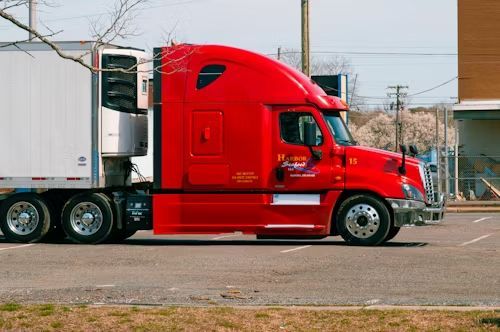 Red semi-truck with a white refrigerated trailer parked outside; sunny day.