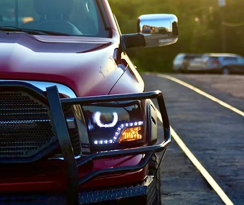 Close-up of a red truck's front end, featuring a black brush guard, custom LED halo headlights, and a chrome side mirror.