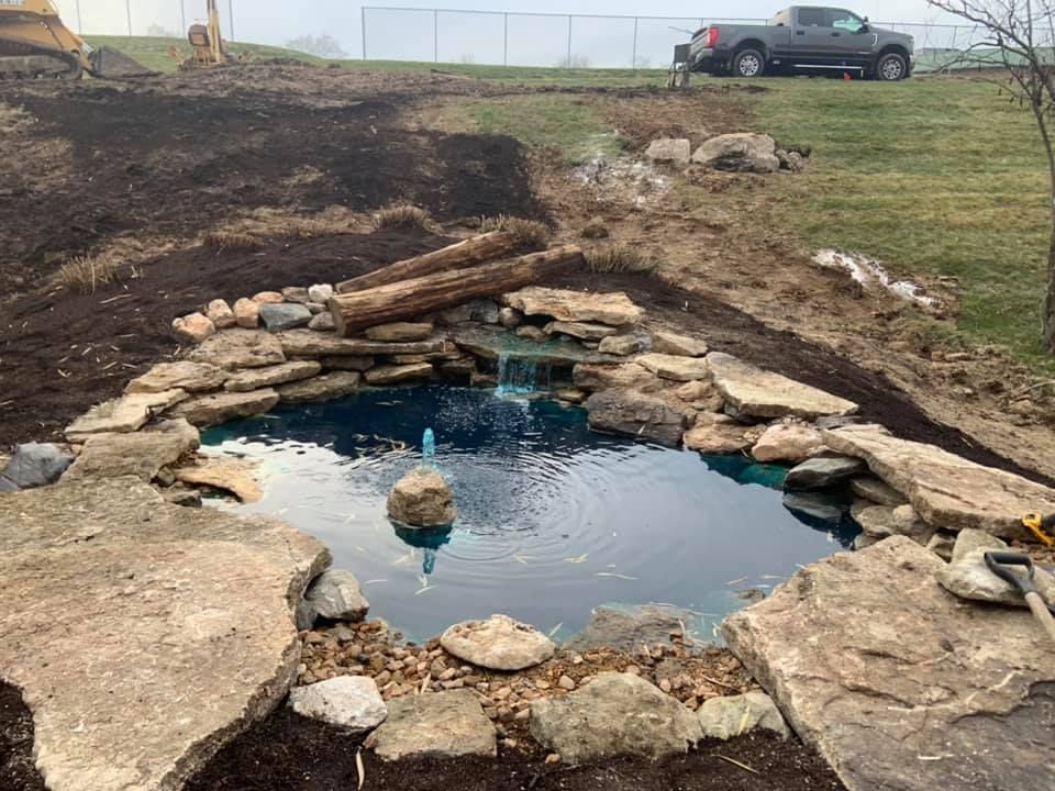 Pond with stone edging, waterfall, and blue water under construction on a grassy hillside.