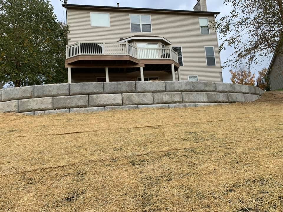 A two-story beige house with a deck sits above a retaining wall on a grassy slope.