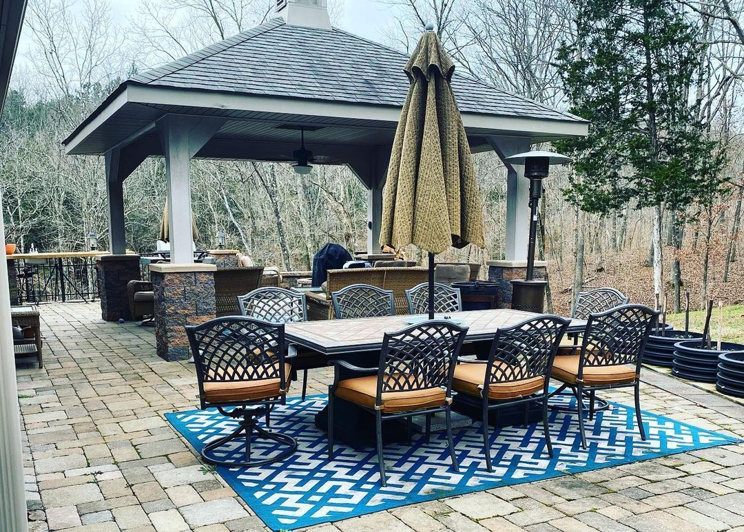 Outdoor patio with gazebo, table, chairs, rug, and umbrella in a wooded setting.