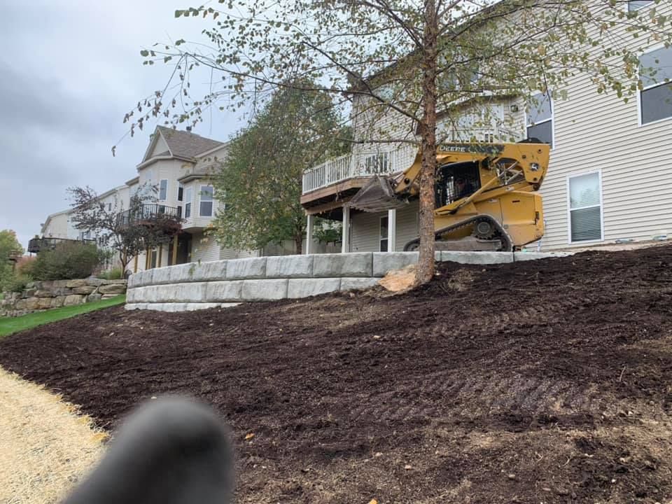 Yellow skid steer next to a house with a retaining wall. Mulched slope in front. Overcast day.