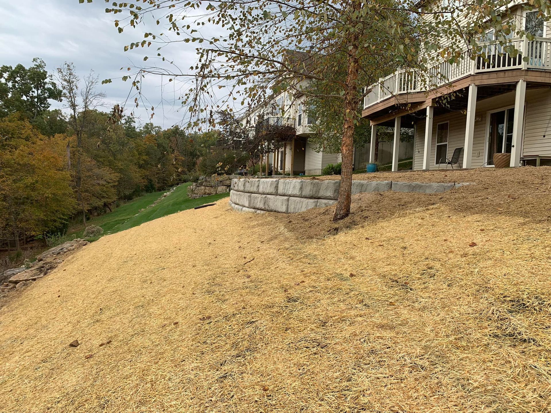 Sloping hillside covered in wood mulch, retaining wall, and house with a deck.