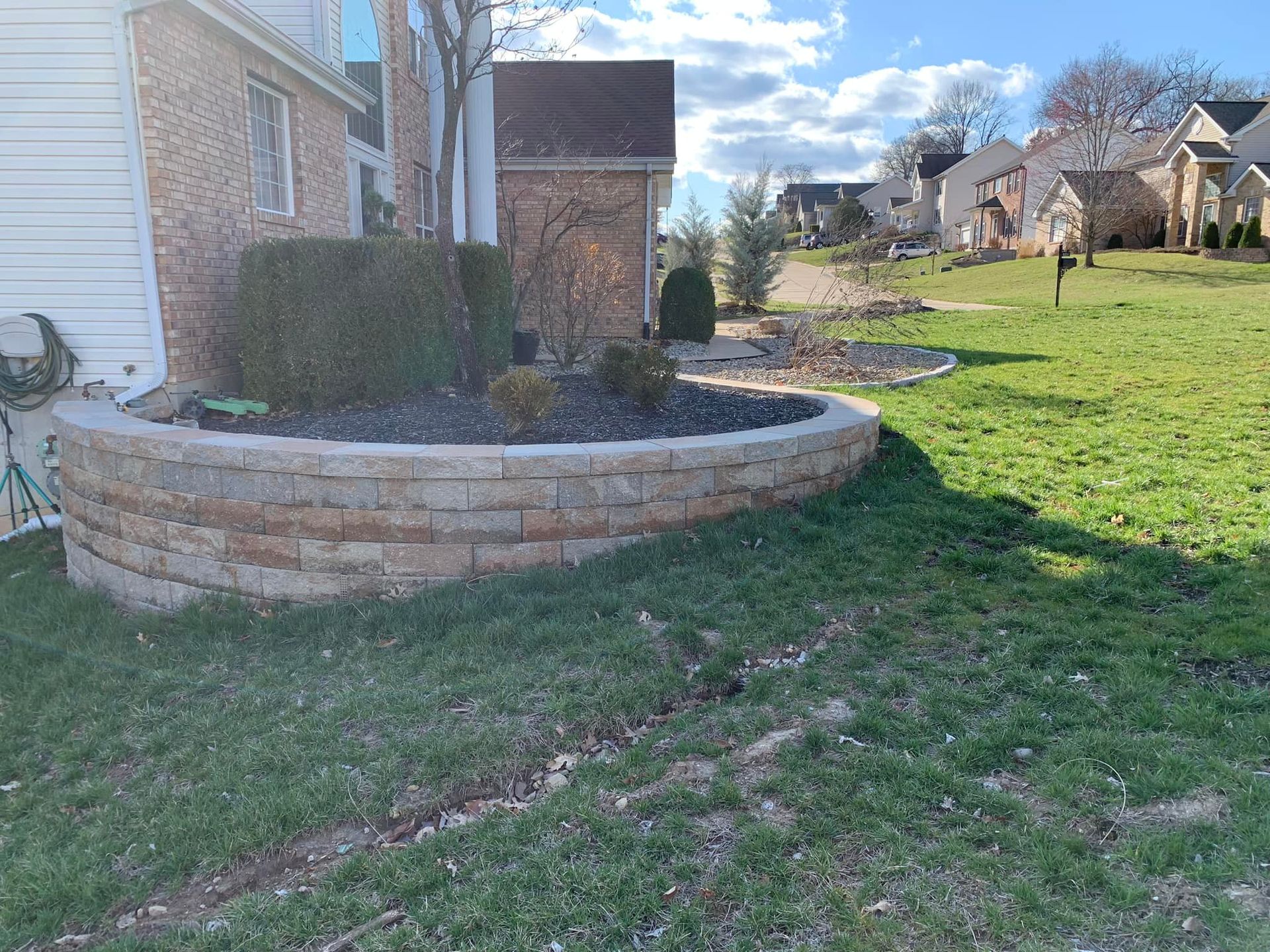 A brick retaining wall borders a garden bed next to a house with a lawn.