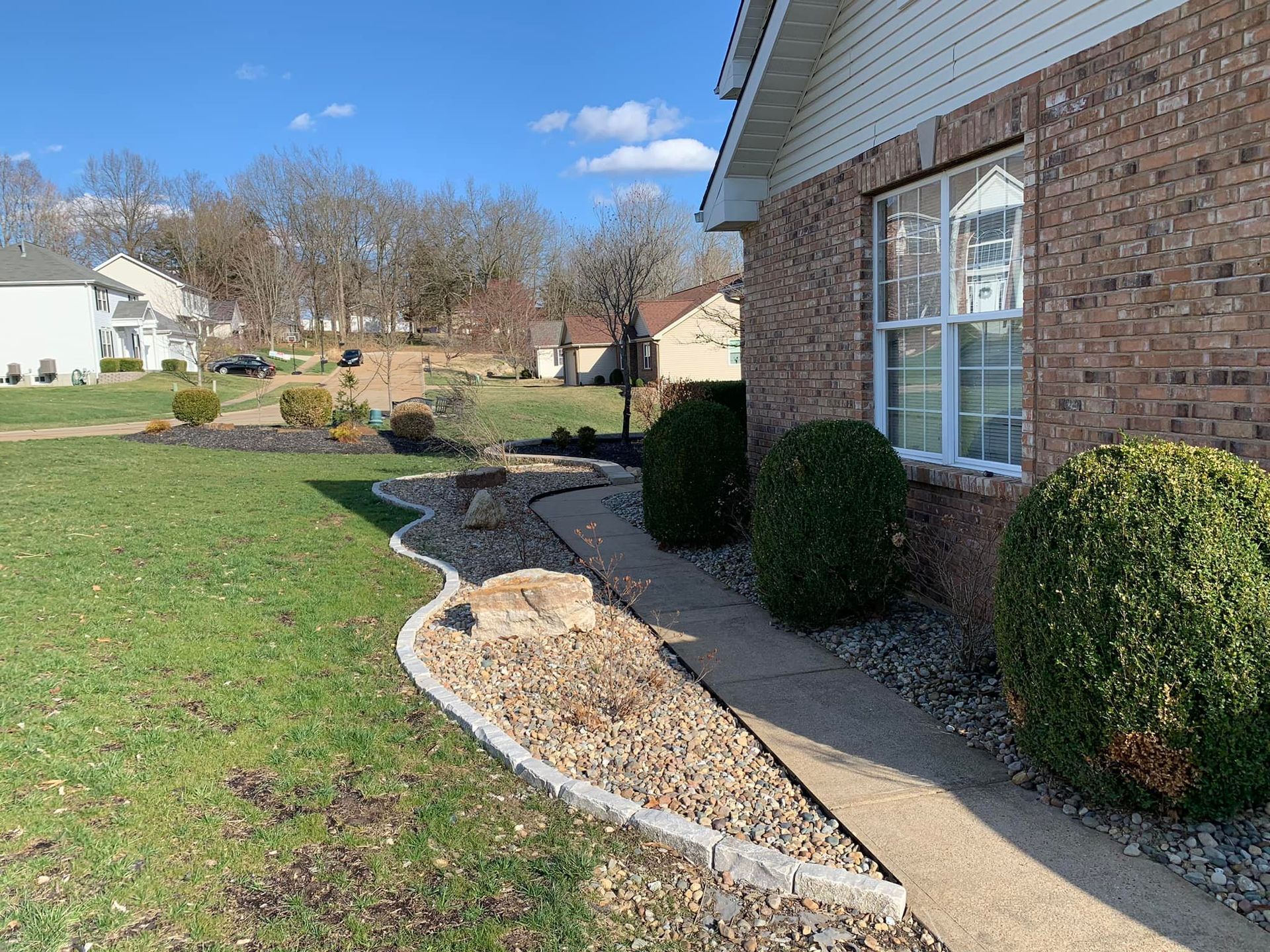 Brick house with green lawn, sidewalk, and landscaped yard with bushes and rocks.