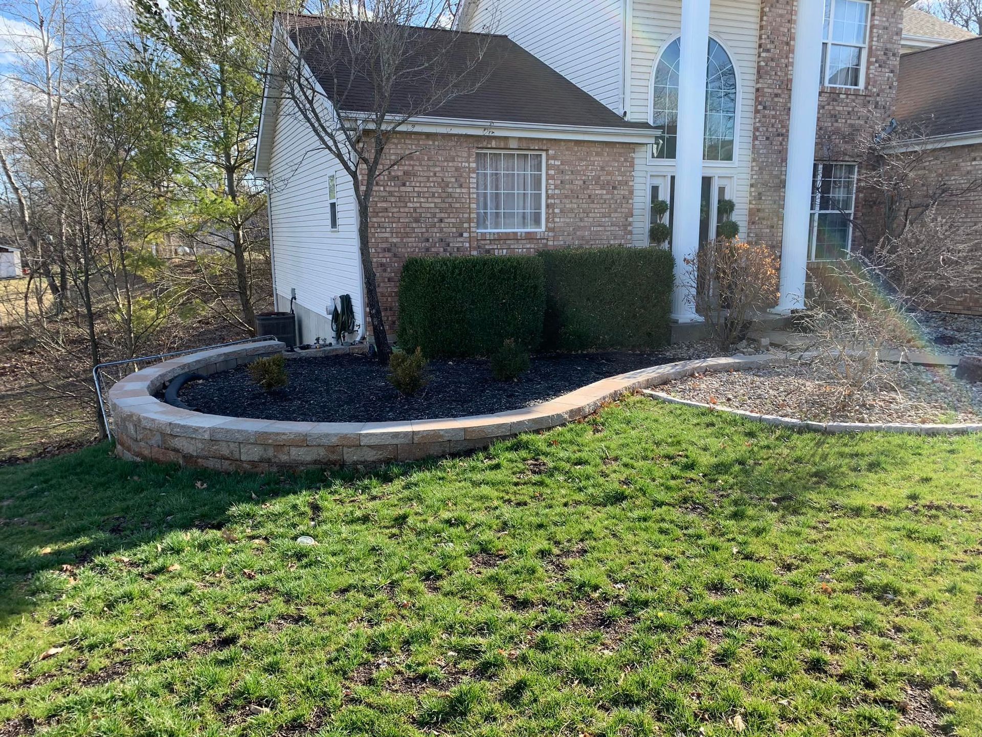 Low retaining wall with dark mulch bed, shrubs, and grassy lawn in front of a house.