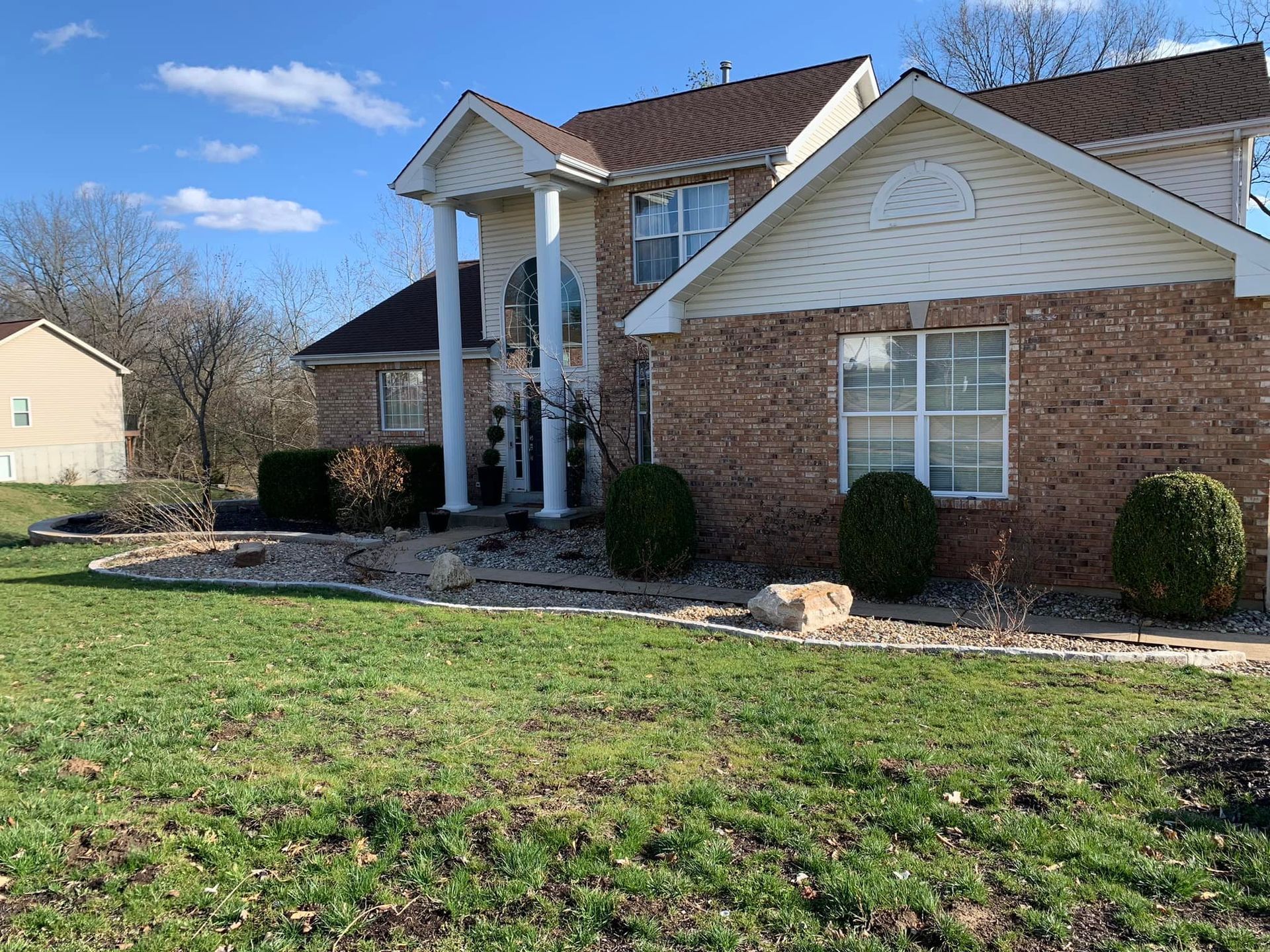 Two-story brick house with white trim, columns, and a landscaped front yard with green grass and shrubbery.