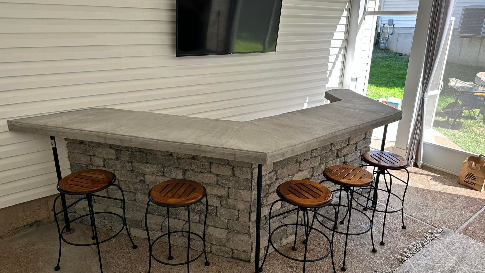 L-shaped outdoor bar with gray stone base, concrete countertop, and wooden stools. TV mounted above.
