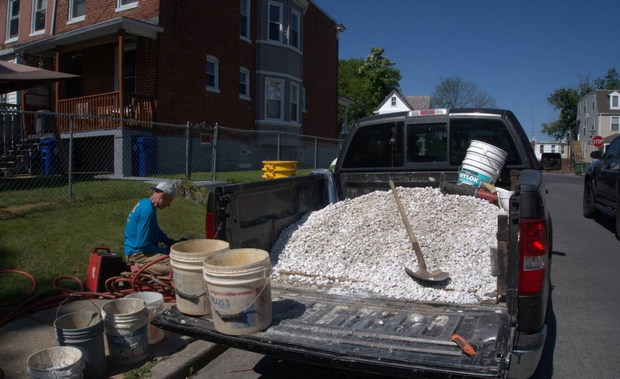 Person working with buckets and gravel in back of pickup truck on a sunny residential street.