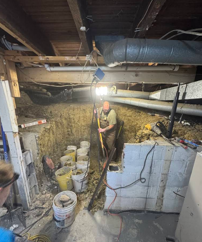Man excavating a trench under a house, buckets of dirt lining the side, indoor setting.