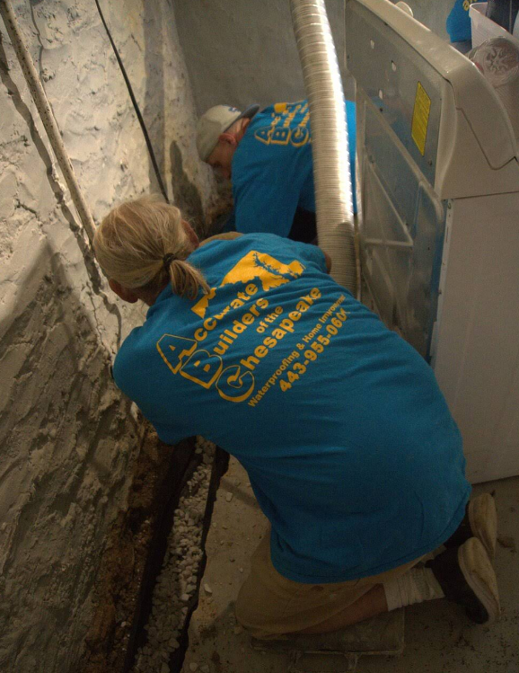 Two people in blue shirts, crouching near a washing machine in a basement, working on something.