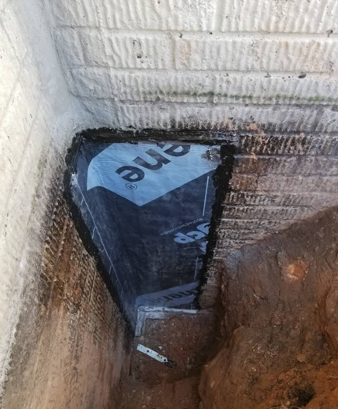 Black tar paper covers a triangular section of a foundation wall. Soil surrounds.
