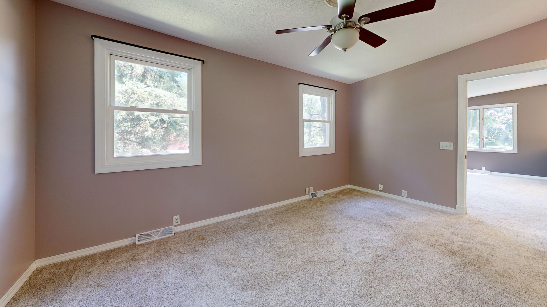 Empty beige bedroom with carpet, two windows, ceiling fan, and doorway to adjoining room