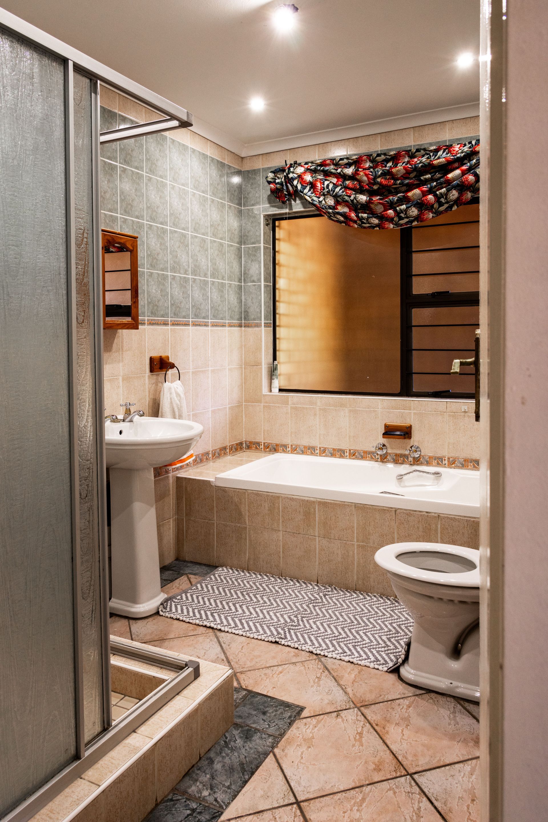 Small bathroom with glass shower, pedestal sink, toilet, and beige tub under a window with red garland.