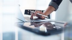 A person is holding a pen in front of a laptop and a tablet.