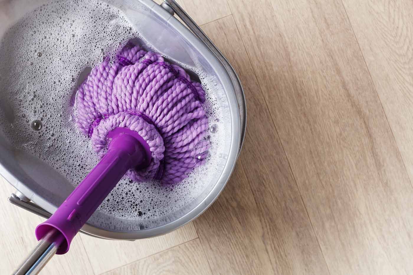 A purple mop is sitting in a bucket of soapy water on a wooden floor.