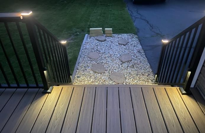 Composite deck steps with built-in lights leading to a gravel path with stepping stones. Black railing. Nighttime.