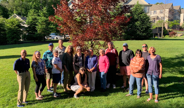 Group of people standing on a grassy lawn in front of trees and houses on a sunny day