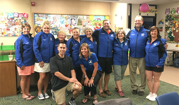 Group of people in blue shirts posing together in a colorful classroom or library.