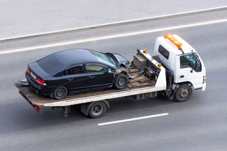 Black car damaged in accident being towed on a flatbed truck on highway.