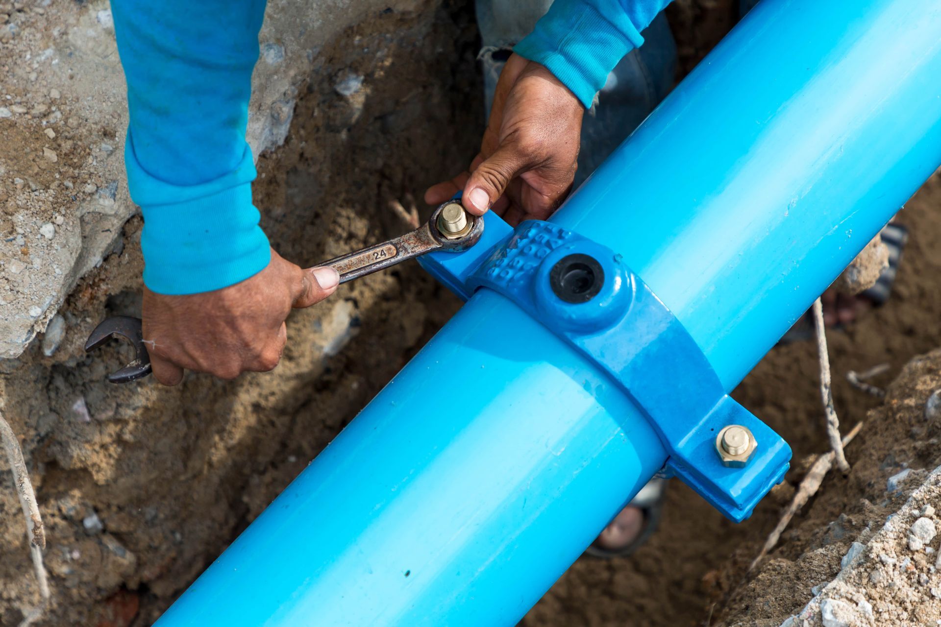 Person using a wrench to tighten bolts on a blue water pipe clamp in a trench.