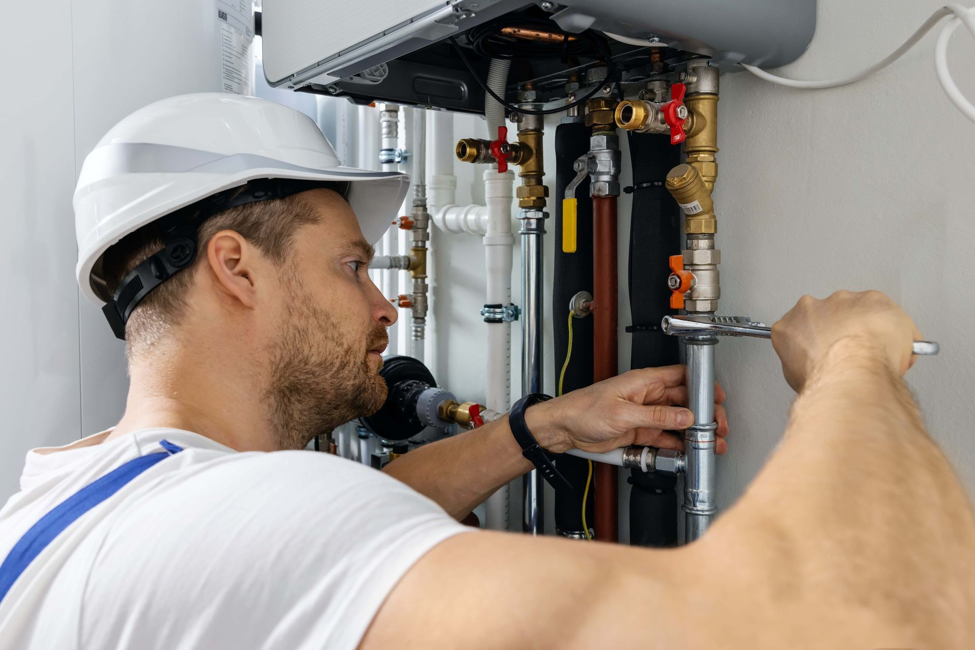 Plumber in a white hardhat, using a wrench on pipes near a boiler.