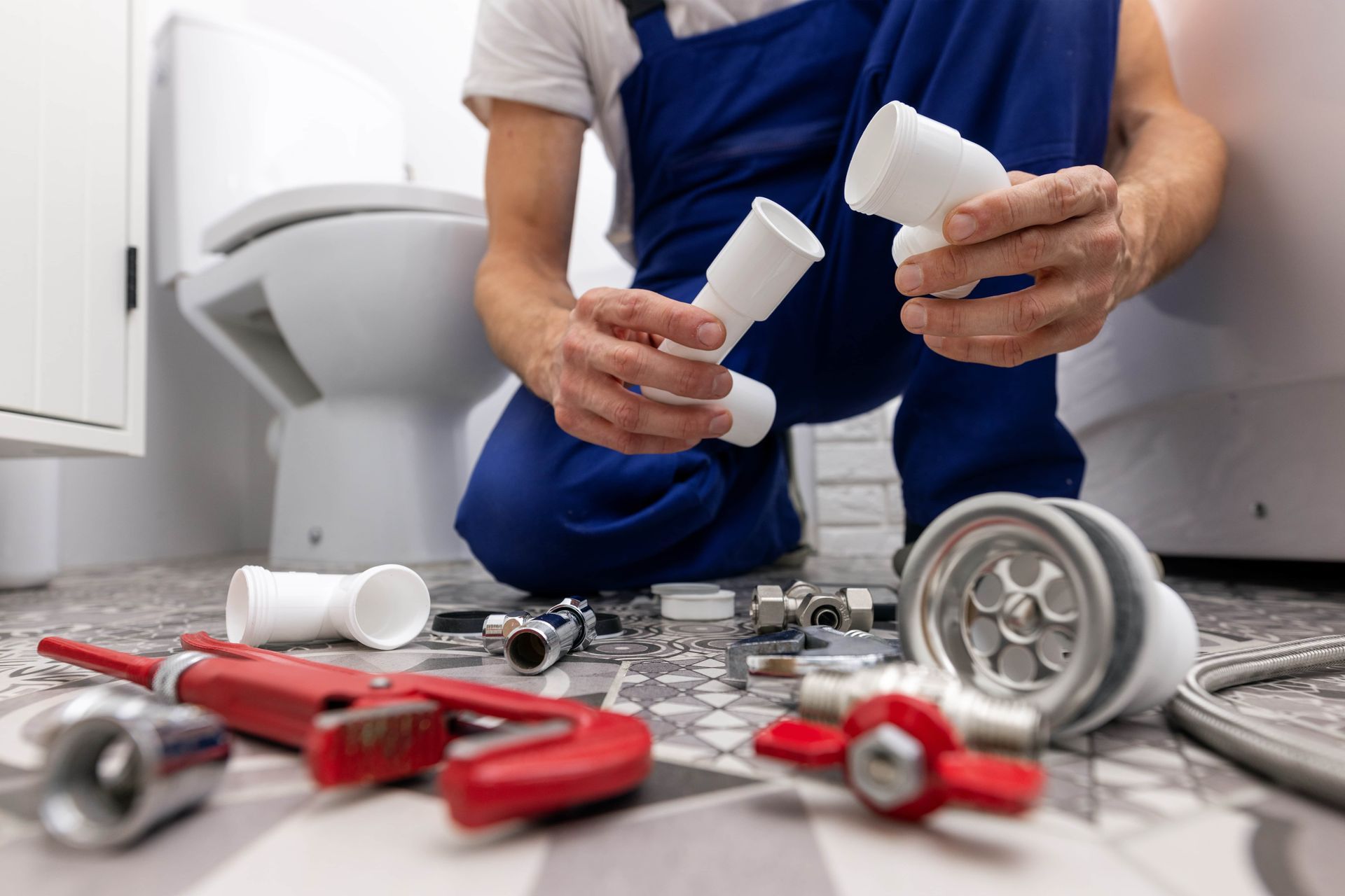 Plumber in blue overalls kneeling in bathroom, assembling plumbing parts near toilet and tools.