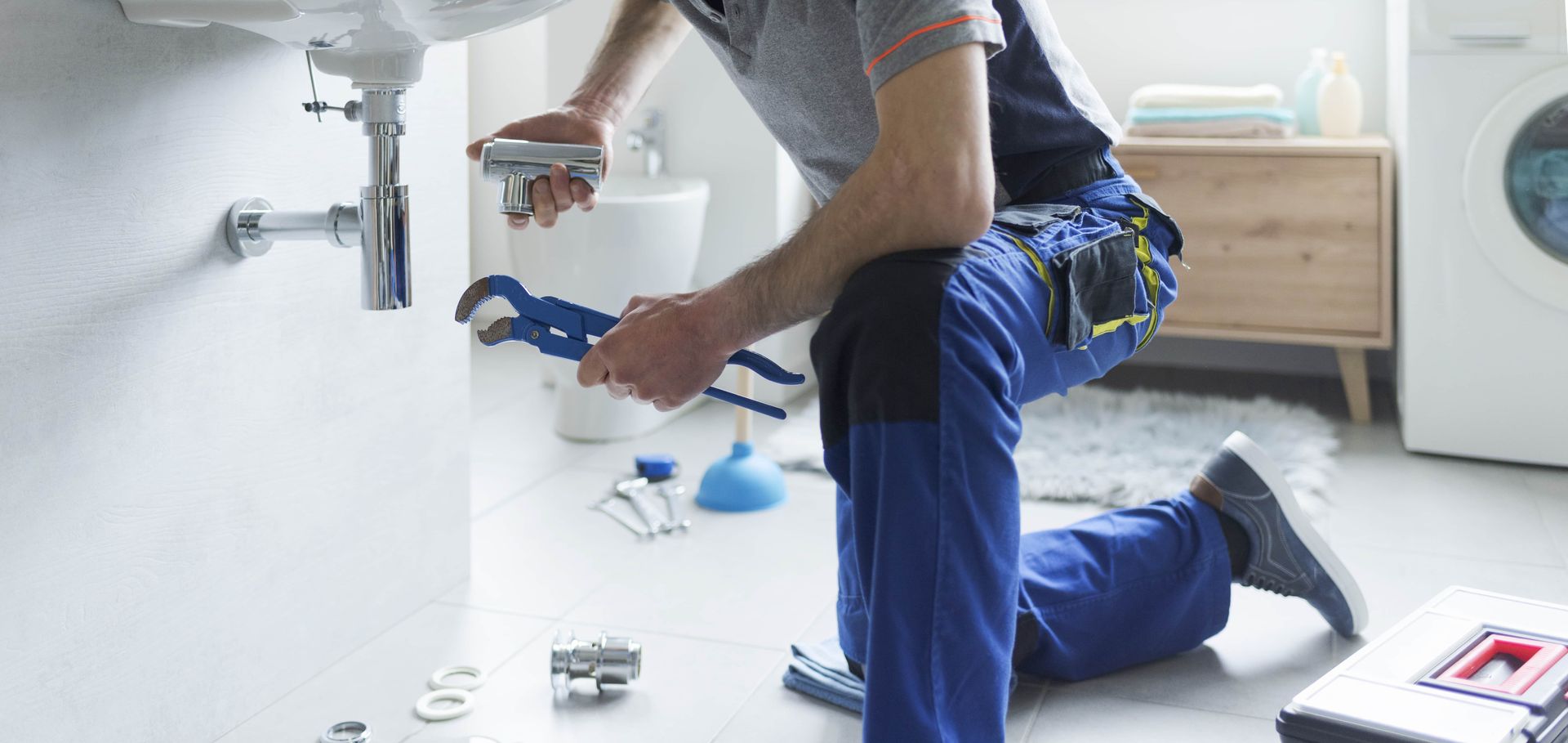 A plumber kneels while using tools to work on a sink in a bathroom.