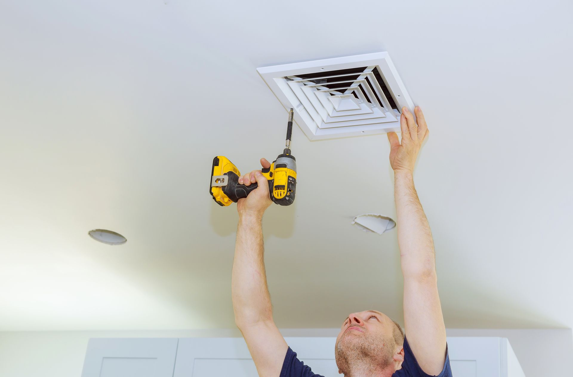 A man is using a drill to install an air vent on the ceiling.