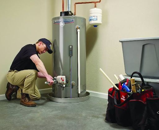 Technician kneels beside a water heater, adjusting a valve in a utility room with tools nearby.
