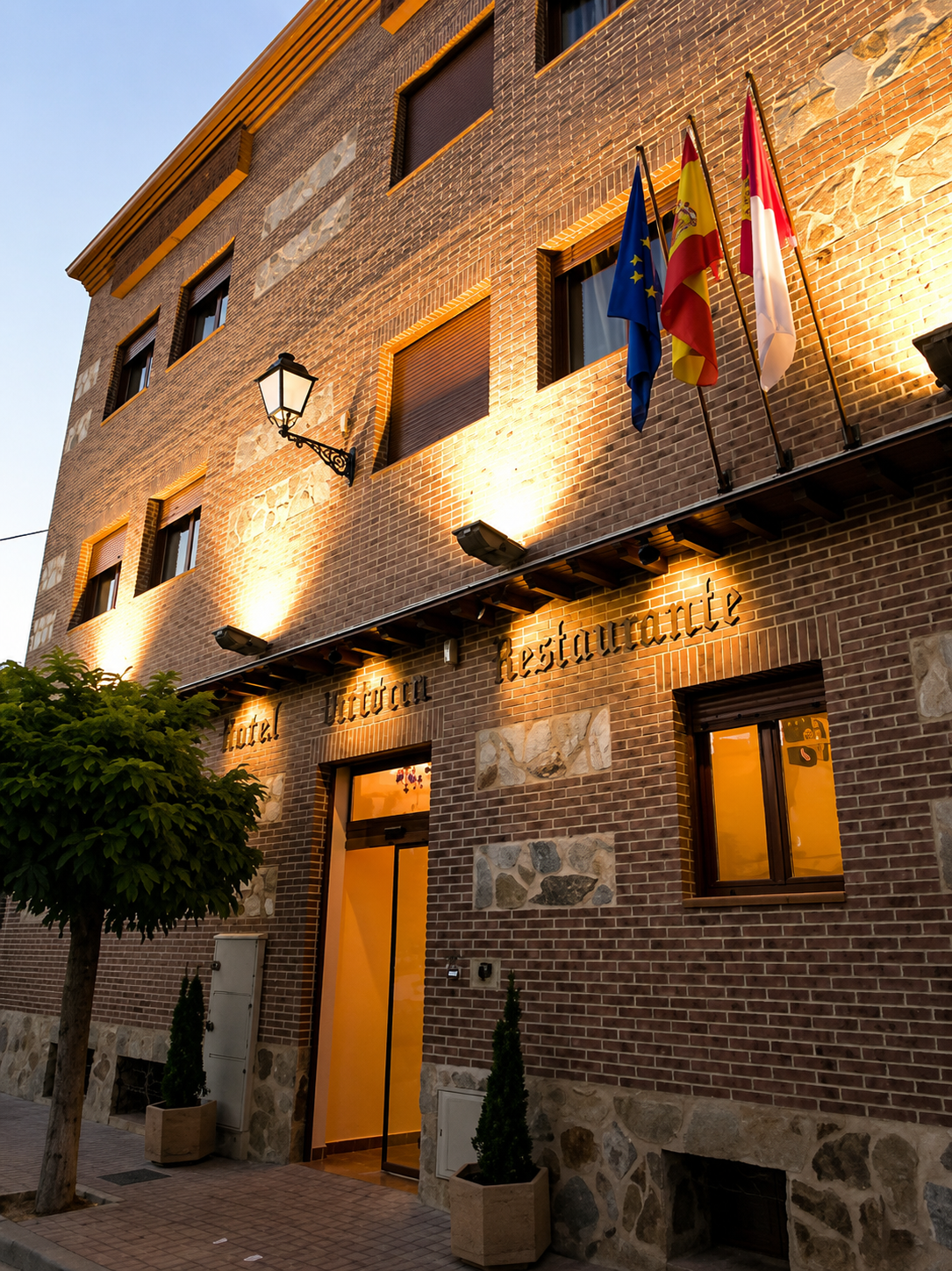 Entrada de hotel de ladrillo iluminada al anochecer con banderas y ventanas con iluminación cálida.