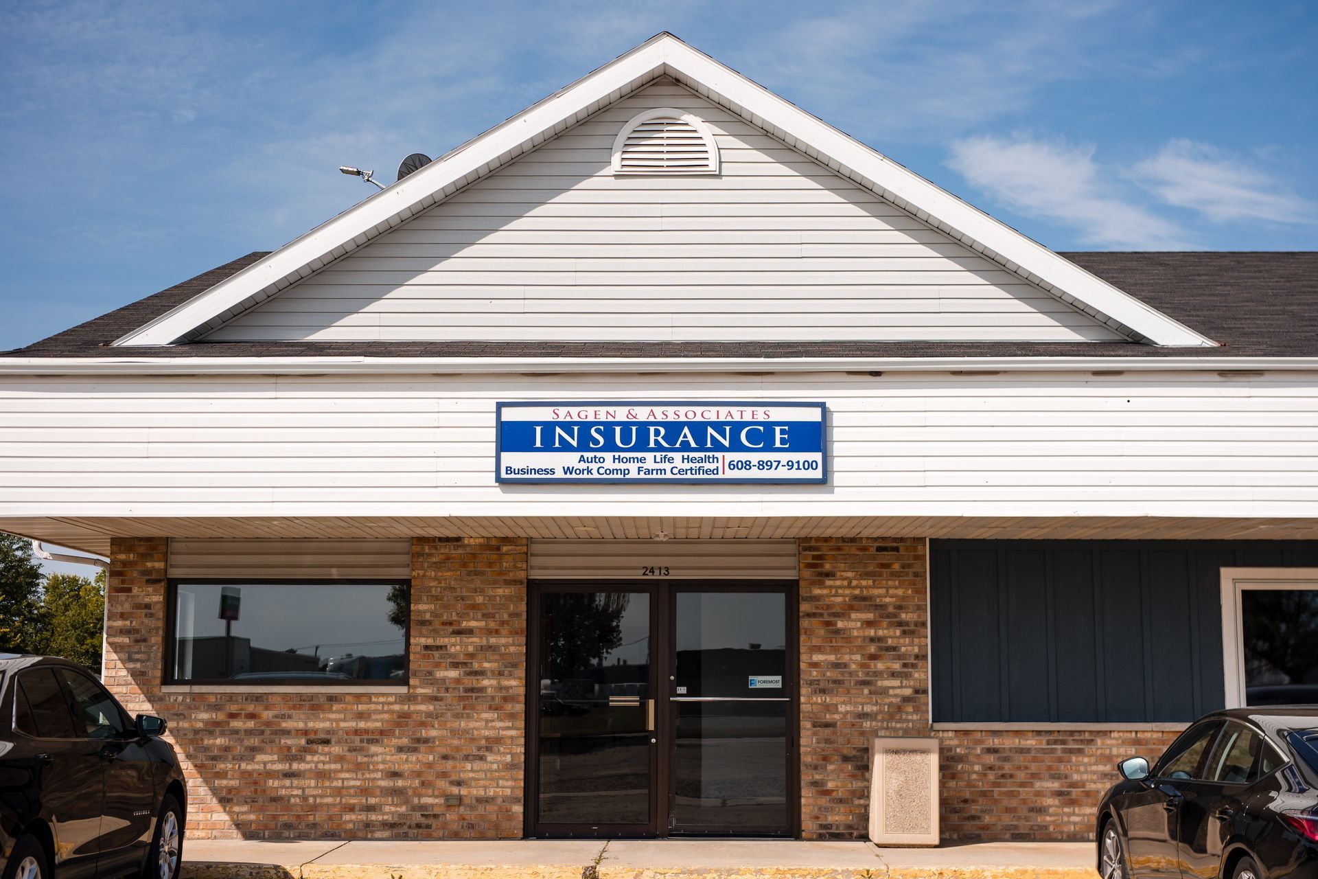 Insurance agency exterior, blue sign above entrance, brick and blue siding, two parked cars.