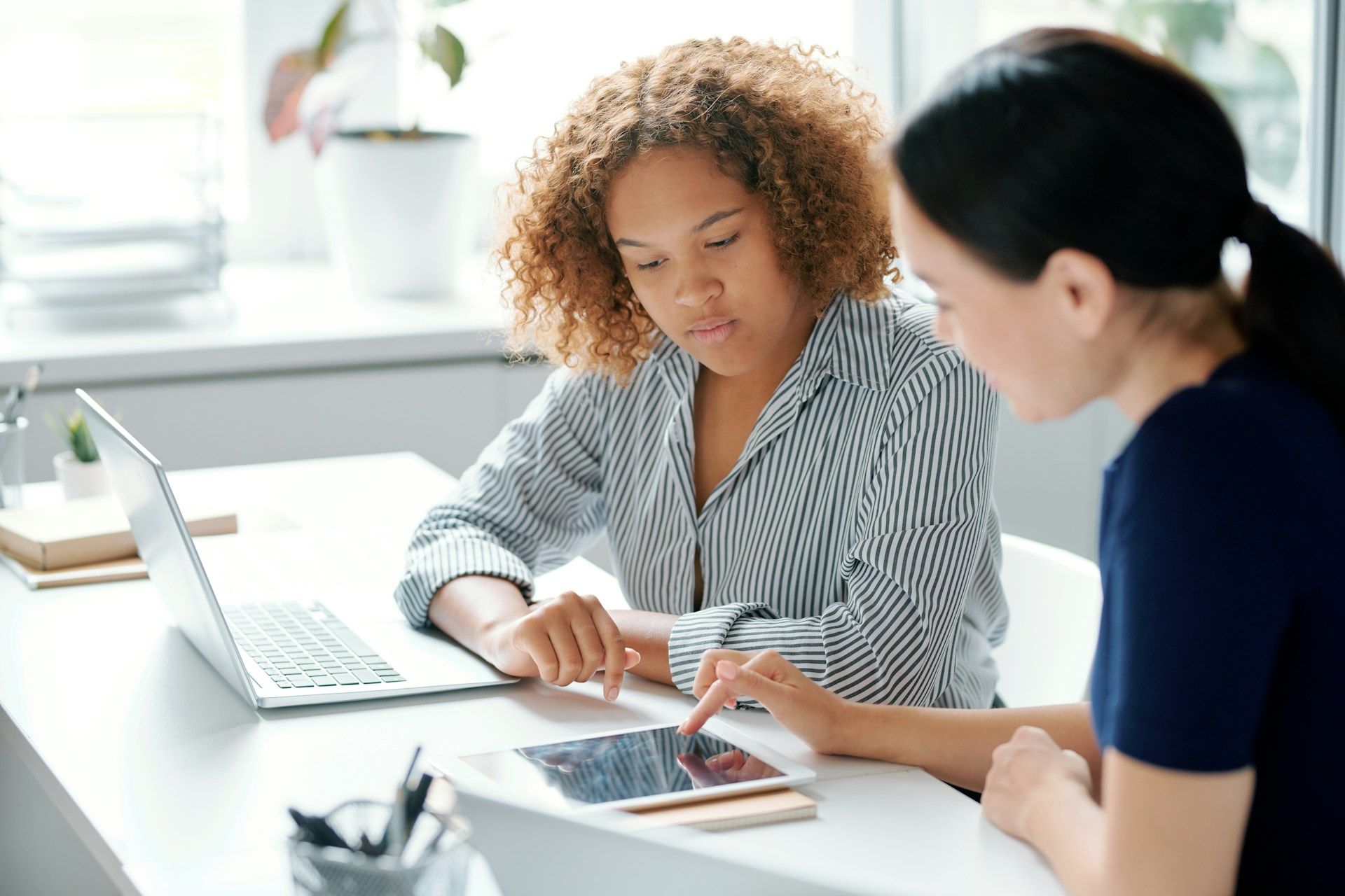 Two women are sitting at a table looking at a tablet and a laptop.