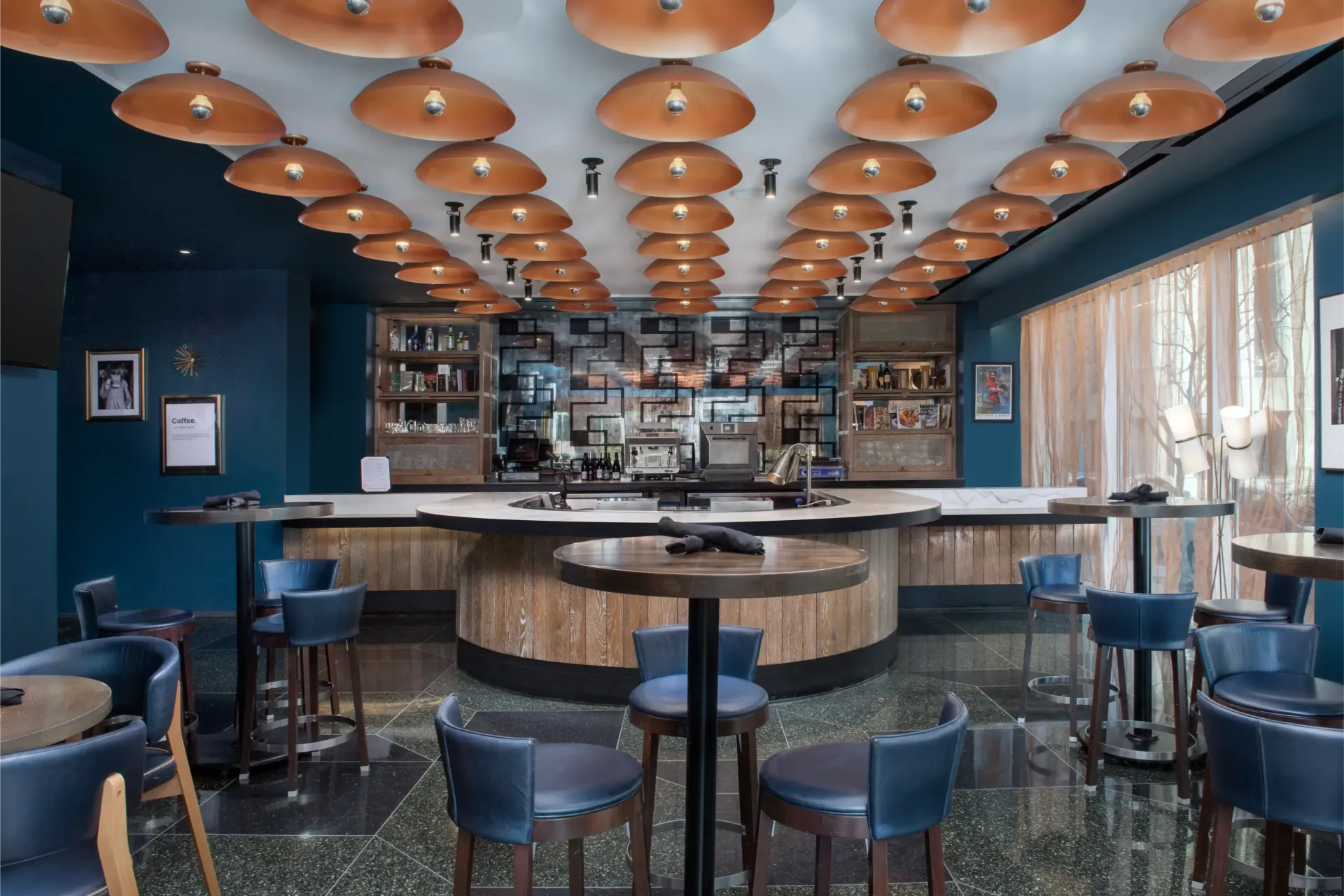 A restaurant with tables and chairs and a bar with copper lights on the ceiling.