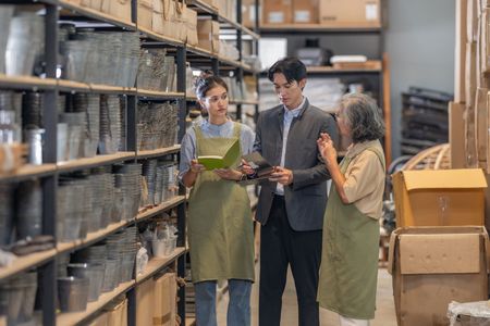 Three people reviewing inventory in a warehouse aisle lined with stacked bins and cardboard boxes